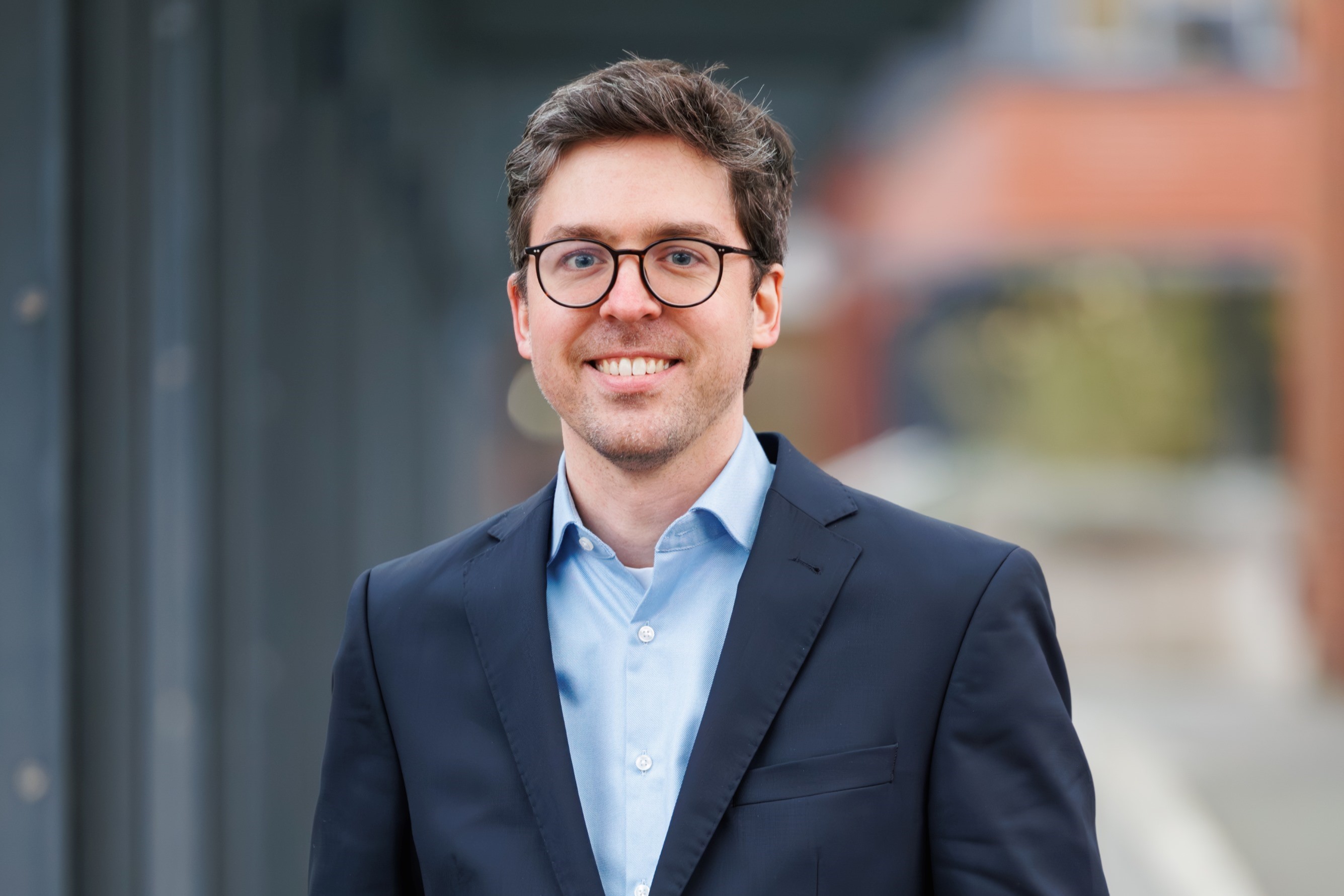 Mann mit Brille und Anzug lächelt in urbaner Umgebung. Im Hintergrund sind unscharfe Gebäude und Bäume zu sehen.