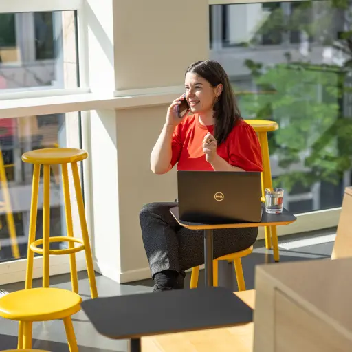 A man sits in a cozy corner, talking on the phone while using a laptop on a wooden table. Soft lighting creates a focused atmosphere.