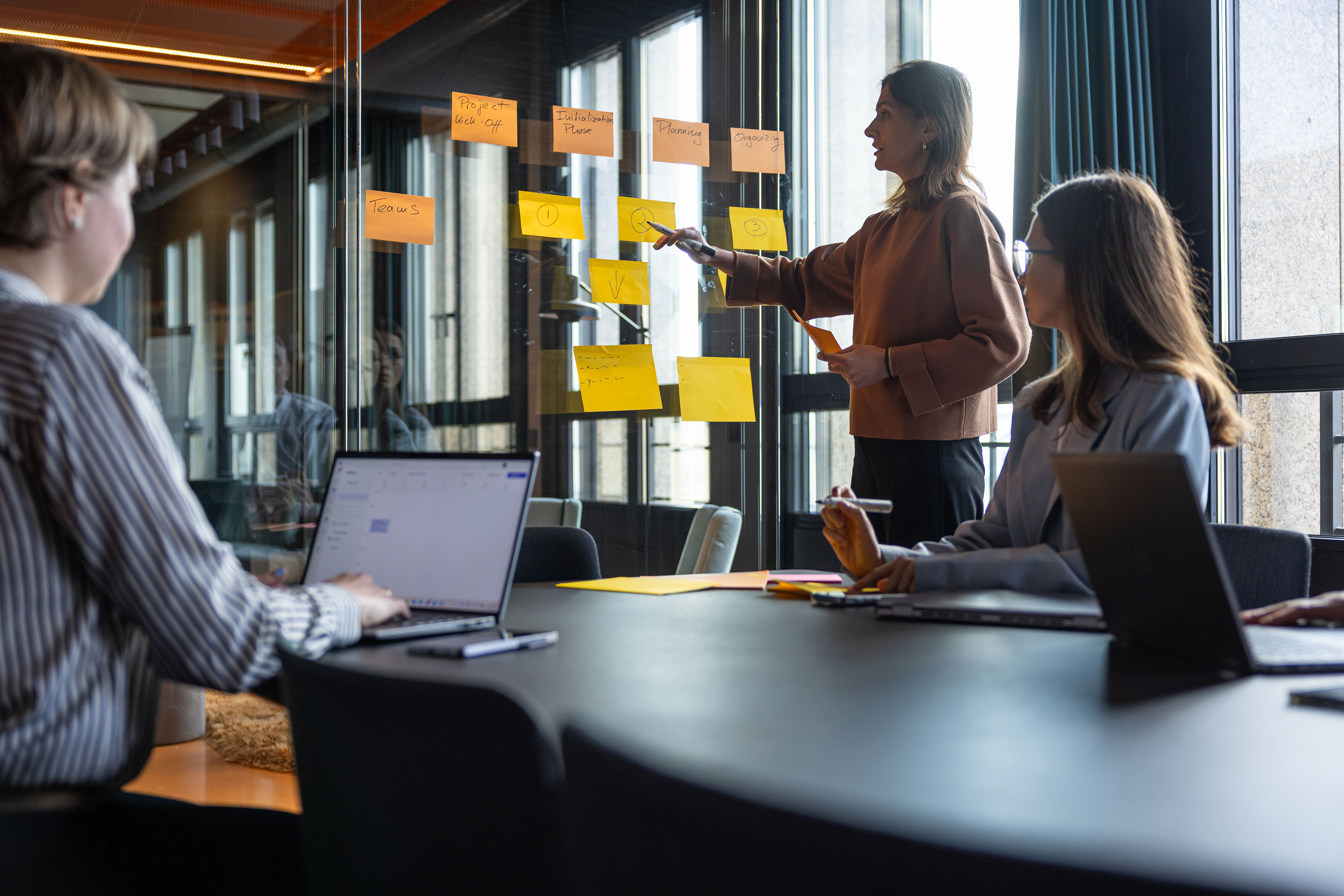 Two women are discussing and organizing colorful Post-it notes on a glass wall in an office setting.