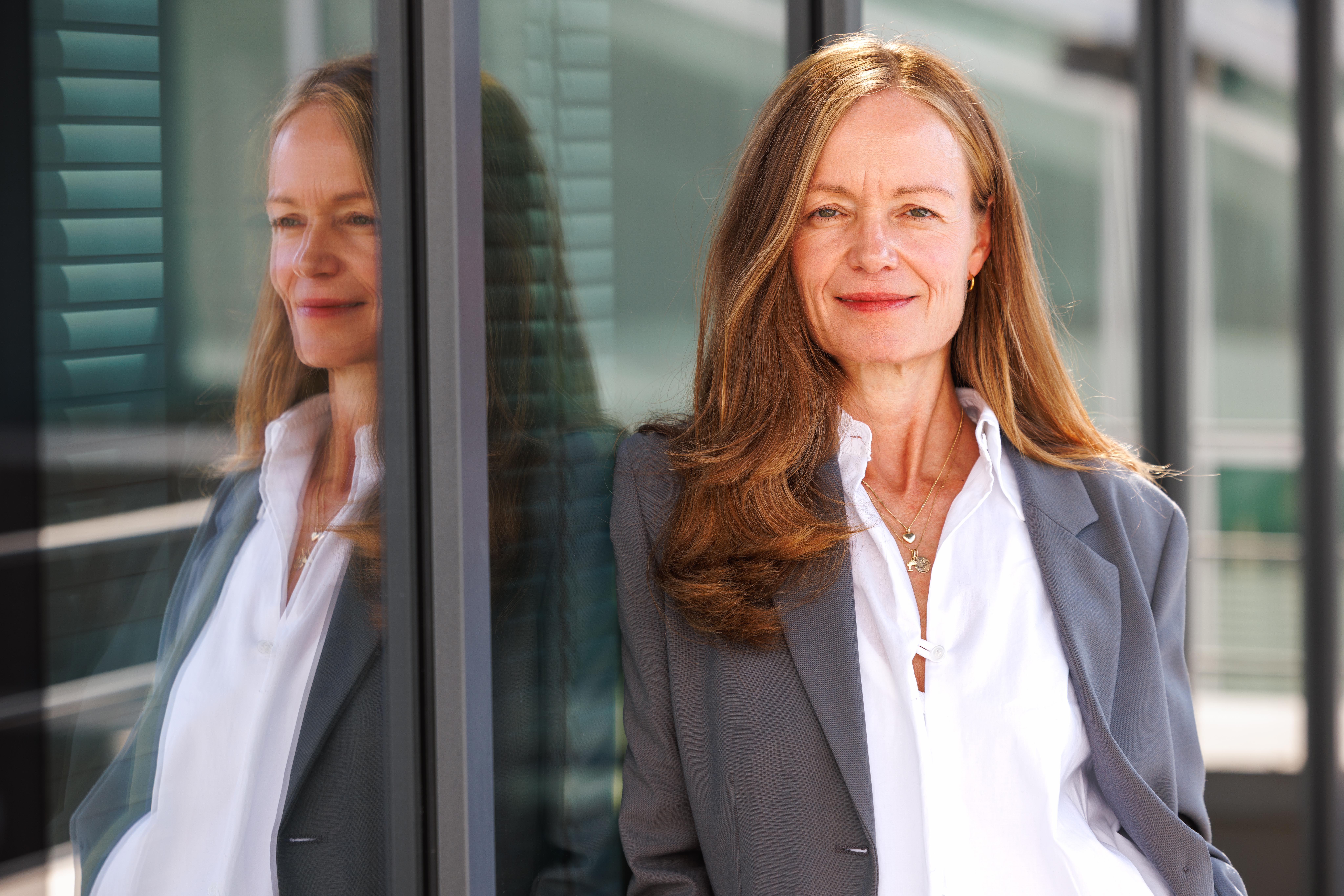 A woman with long blonde hair smiles while leaning against a metal structure. She wears a grey jacket and stands outdoors.